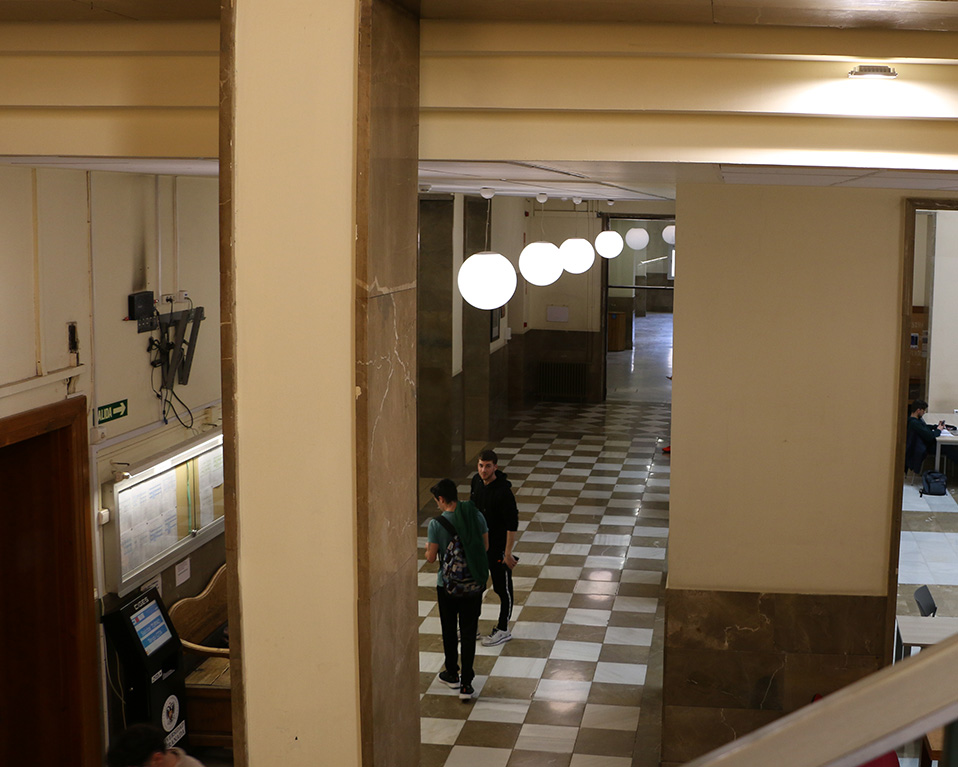 Dos alumnos en un pasillo vistos desde las escaleras en la Facultad de Ciencias Políticas y Sociología. A la derecha, otros dos alumnos estudiando.