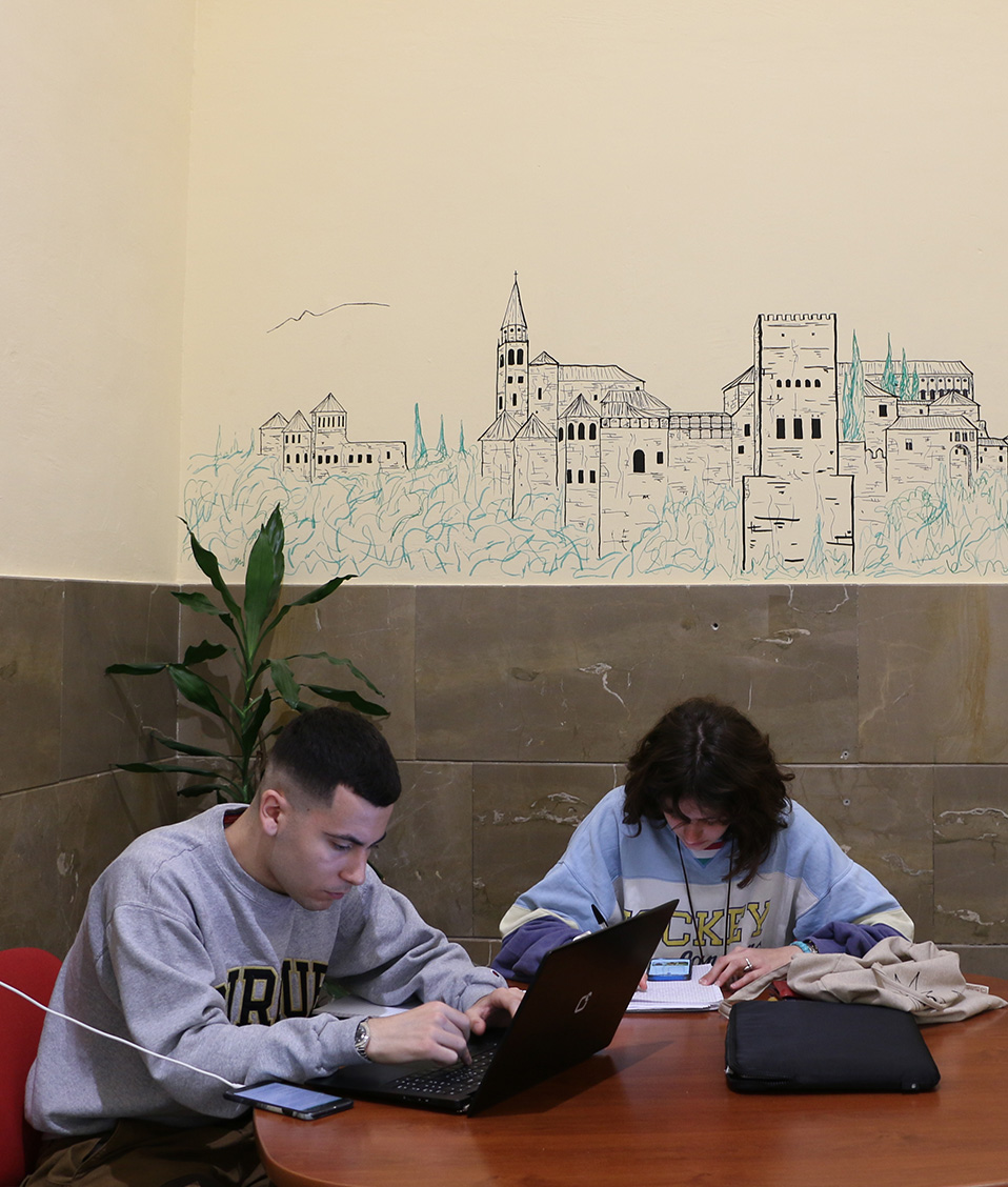Pasillos de la Facultad de Ciencias Políticas y Sociología. Dos alumnos sentados a una mesa estudiando. Dibujo de la Alhambra al fondo.